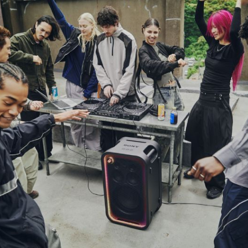 Grupo de jóvenes reunidos en una terraza al aire libre bailando alrededor de una mesa de DJ donde se encuentra una bocina de torre Sony ULT TOWER con luces encendidas alrededor de la rejilla.