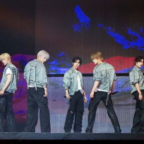 Los cinco integrantes del grupo de K-pop TOMORROW X TOGETHER posan de pie sobre un escenario de concierto vestidos con chaquetas grises estilo industrial frente a pantallas gigantes de fondo.   