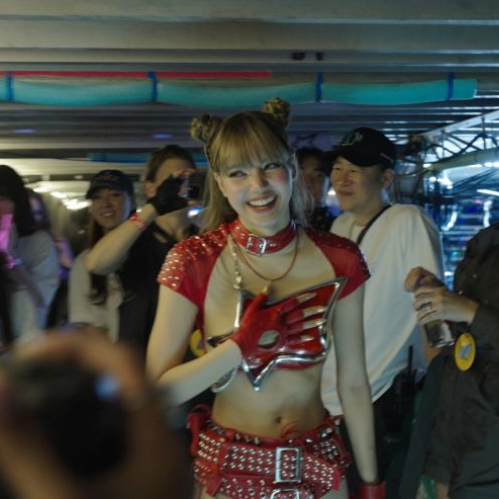 Fotograma de la película "Always Lalisa". En el centro se observa a la cantante LISA sonriendo tras bambalinas, peinada con dos rodetes superiores y flequillo. Luce un atuendo de escenario rojo y plateado con aplicaciones metálicas en el torso, gargantilla de cuero roja y guante rojo. A su alrededor se encuentra su equipo de trabajo sonriendo y aplaudiendo dentro de la estructura de un escenario.