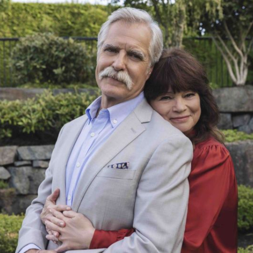 Los actores Henry Czerny y Valerie Bertinelli posan juntos al aire libre en un jardín rodeado de vegetación y muros de piedra. Henry Czerny, al frente, luce cabello canoso y un bigote blanco, viste un saco gris claro, camisa azul y un pañuelo de bolsillo estampado. Valerie Bertinelli lo abraza cariñosamente por la espalda rodeándole el pecho con los brazos, vistiendo una blusa roja de manga larga y sonriendo de forma serena hacia la cámara.
