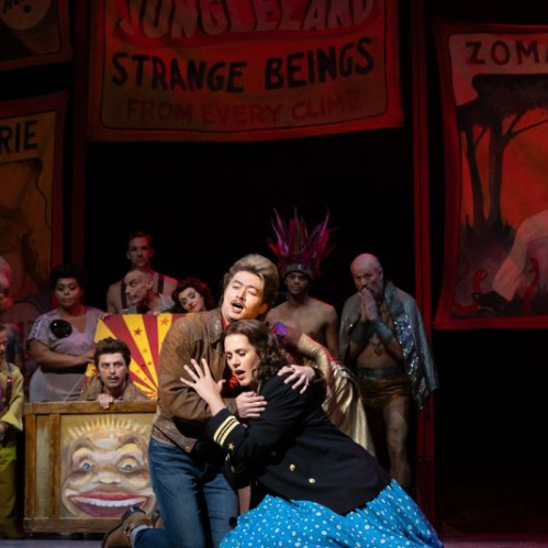 Escena dramática del reestreno de Così fan tutte ambientado en Coney Island con cantantes de ópera abrazados frente a letreros vintage de feria y artistas circenses.
