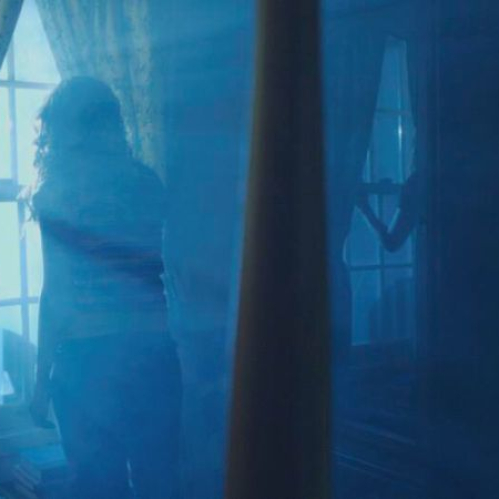 Fotografía promocional en plano medio en interiores con un tono azulado y brumoso. Una persona de espaldas con cabello largo se encuentra frente a una ventana con cortinas claras, mirando hacia el exterior. A la derecha se aprecia la silueta de un mueble de madera reflejado y una pared oscura en un ambiente envuelto en humo o neblina azul.