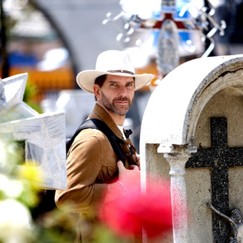 Arap Bethke posa en primer plano luciendo un sombrero de ala ancha color beige y una chaqueta de gamuza marrón, sujetando las correas de una mochila. Al fondo, fuera de foco, se observan tumbas, cruces de mármol y flores en un panteón tradicional mexicano.