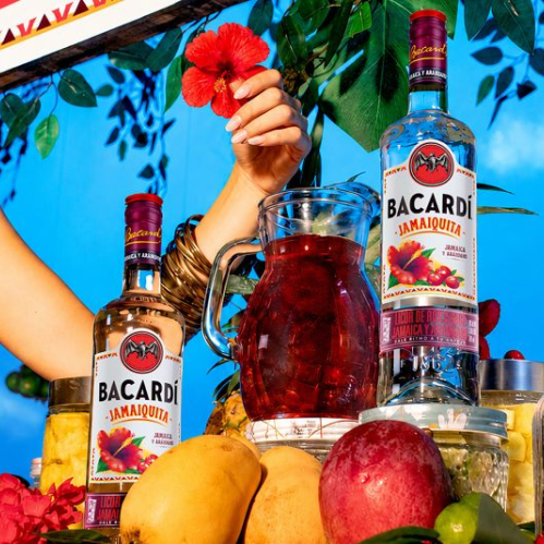 Botellas de cristal del ron saborizado Bacardí Jamaiquita expuestas en un puesto de frutas con flores rojas de jamaica, mangos y piñas. Al centro destaca una jarra de cristal con la bebida preparada de color rojo oscuro sobre un fondo de vegetación y cielo azul.