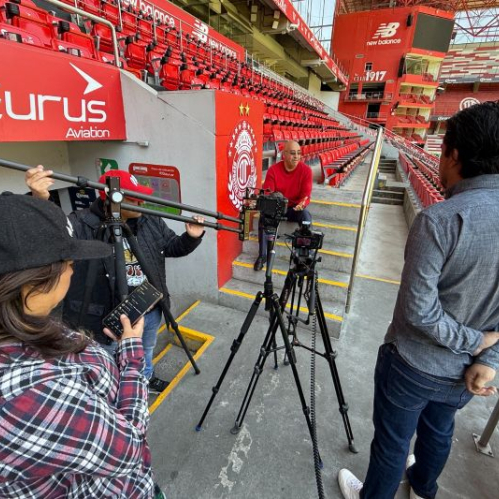 Equipo de producción grabando una entrevista en un estadio con cámaras, micrófonos y personal técnico.