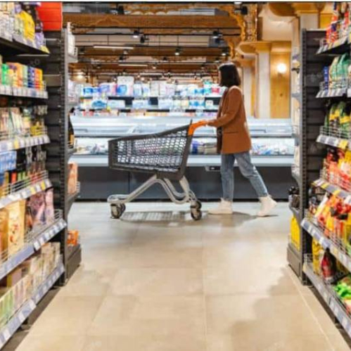 Fotografía de una persona empujando un carrito por un pasillo de supermercado con estantes llenos de productos empaquetados y una zona refrigerada al fondo.