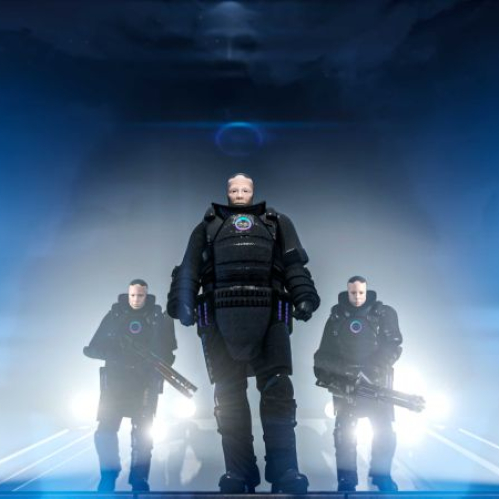 Tres personajes con armaduras futuristas posan en un entorno iluminado por luces azules y humo, evocando una escena de acción.