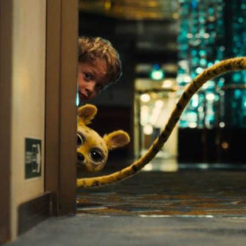 Fotografía cinematográfica horizontal en formato panorámico que muestra un pasillo interior de un barco de crucero con moqueta azul decorada e iluminación cálida de fondo. En primer plano, asomándose desde detrás de un marco de puerta en el lado izquierdo, se observa a un niño pequeño junto a un bebé Marsupilami amarillo con manchas negras y grandes ojos oscuros. Su larga cola amarilla y negra acentuada con manchas se extiende a lo largo del pasillo.