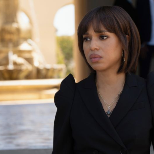 Retrato en plano medio de la actriz Kerry Washington interpretando a su personaje Eleanor. Luce un peinado estilo bob con fleco recto, viste un saco negro de vestir con escote cruzado y un collar dorado con un dije sutil. Mantiene una expresión seria y contemplativa mirando ligeramente fuera de cámara, con una fuente de agua y la entrada de una residencia iluminada al fondo en tonos cálidos fuera de foco.
