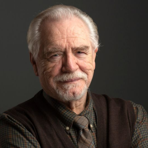 Retrato de Brian Cox con los brazos cruzados, vistiendo camisa y chaleco, sobre fondo oscuro.