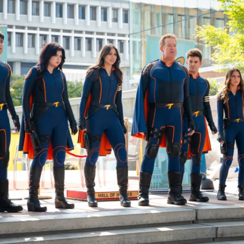 Fotografía de estudio en plano entero del elenco principal de la película "La Furia de los Thundermans". De izquierda a derecha, siete miembros del reparto (Diego Velázquez, Ariel Winter, Kira Kosarin, Chris Tallman, Jack Griffo, Addison Riecke y Rosa Blasi) visten trajes tácticos ajustados de superhéroes en azul marino con detalles en naranja e insignias con la letra 'T' en el pecho y cinturones, posando alineados al aire libre frente a una pancarta del 'Hall of Heroes'.