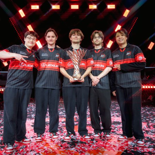 Los integrantes de 100 Thieves vistiendo sus camisetas rojas y negras posan sonrientes en el escenario sosteniendo el trofeo de campeones del VCT Americas Stage 2 mientras cae confeti rojo y blanco.