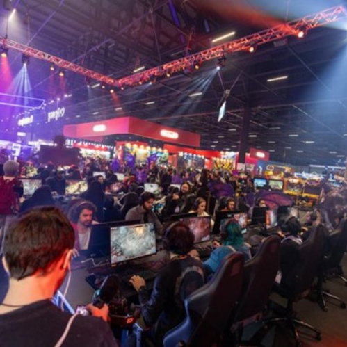 Área de un evento gamer con estaciones de juego, público participando y luces de escenario en tonos azules y rojos.