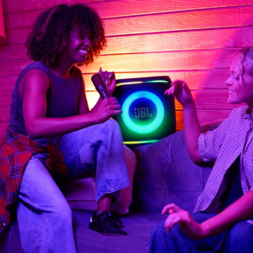 Fotografía de dos personas en un ambiente de entretenimiento interior mientras interactúan con un altavoz JBL iluminado.