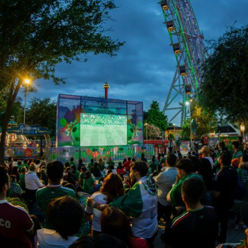 Multitud reunida en un espacio público observando un partido de futbol en pantalla gigante.