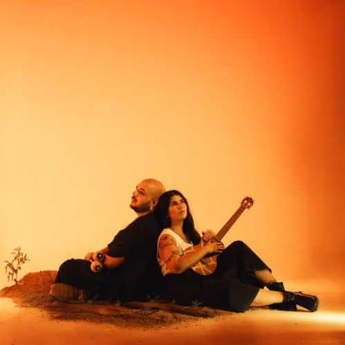 Dos artistas sentados espalda con espalda sobre un pequeño montículo de tierra, cada uno sosteniendo un instrumento musical, iluminados por un fondo cálido en tonos naranjas.