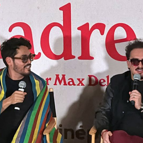 Los comediantes Hugo "El Cojo Feliz" y Ricardo Pérez sentados durante una conferencia de prensa de la película A Toda Madre. El Cojo Feliz viste una prenda multicolor tipo sarape y lentes oscuros sosteniendo un micrófono, mientras que Ricardo Pérez luce lentes oscuros y una chamarra negra conversando con el micrófono. Detrás se observa la escenografía con el título "Madre" en letras rojas.