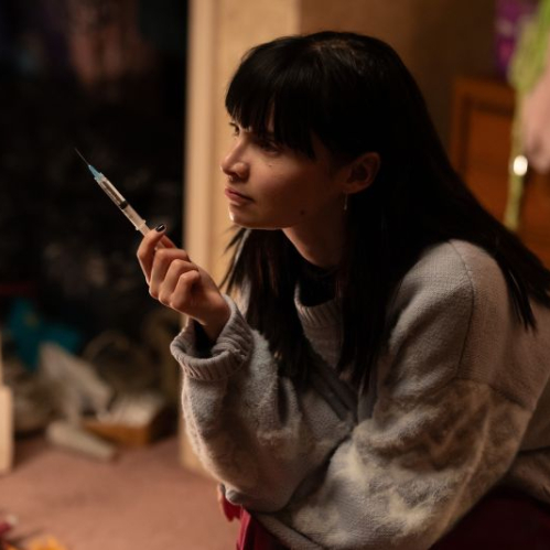 Fotografía de escena en plano medio de Lola Petticrew personificando a Catherine en la serie Furia. La actriz aparece de perfil sosteniendo con cautela una jeringa transparente con líquido entre los dedos. Viste un suéter de lana tejido en tonos gris y blanco, tiene cabello negro con fleco y observa fijamente hacia un costado dentro de una habitación iluminada de forma cálida.