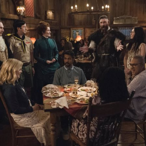 Fotografía de escena en plano general ambientada en un comedor de madera rústica iluminado por lámparas colgantes. Alrededor de una mesa servida con platos y copas de vino, Sam (Rose McIver) y Jay (Utkarsh Ambudkar) escuchan atentamente la conversación del grupo. De pie alrededor de ellos se encuentran varios espíritus con vestimentas de distintas épocas históricas, incluyendo al vikingo Thorfinn haciendo un gesto explicativo con los brazos abiertos.