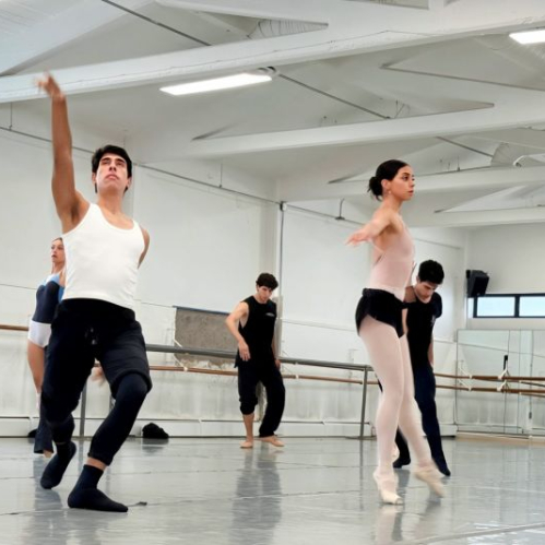 Fotografía de ensayo en salón de danza iluminado donde varios bailarines y bailarinas de la Compañía Nacional de Danza realizan movimientos contemporáneos y clásicos; en primer plano un bailarín con camiseta blanca extiende el brazo hacia arriba mientras una bailarina en puntas realiza una postura grácil.