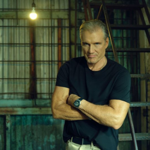 Retrato en plano medio del actor Dolph Lundgren vistiendo una playera negra y pantalón claro, parado con los brazos cruzados frente a una escalera metálica industrial dentro de una estructura de apariencia fabril con una bombilla encendida al fondo.
