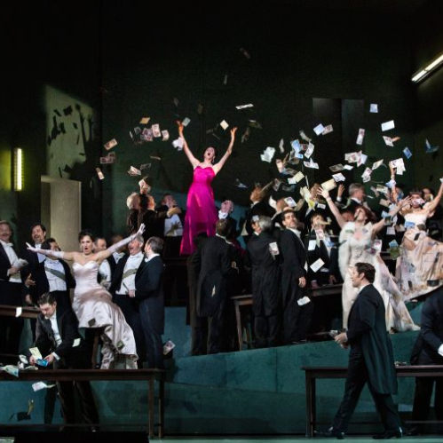 Fotografía de la puesta en escena de la ópera Manon sobre el escenario del Metropolitan Opera House. En el centro, una solista con vestido de gala rosa celebra mientras billetes de papel caen sobre el elenco distribuido en una escenografía inclinada de tonos verdes y elegantes trajes de etiqueta.