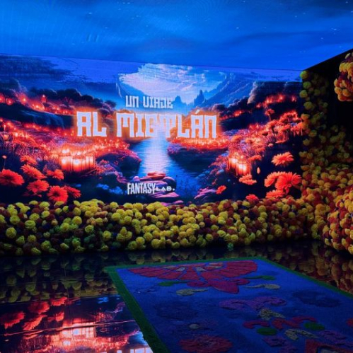 Fotografía del interior de la experiencia inmersiva Un Viaje al Mictlán con un tapete floral al centro en el suelo reflejante, paredes cubiertas de flores de cempasúchil amarillas y naranjas, y una pantalla de fondo que muestra el título de la experiencia sobre un paisaje místico iluminado.