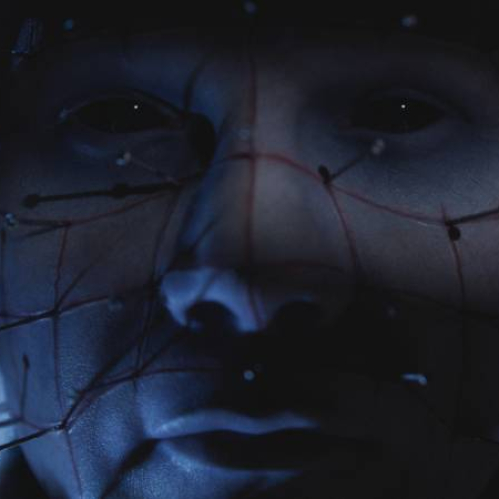 Primer plano de un rostro intervenido con múltiples agujas conectadas por hilos tensos, iluminado por una luz tenue que resalta las sombras y acentúa la atmósfera inquietante.