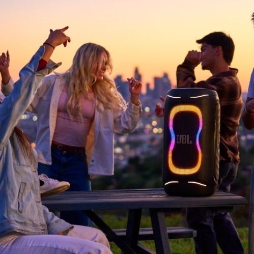 Grupo de jóvenes bailando y disfrutando al aire libre al atardecer en una terraza con vista a la ciudad, junto a una bocina portátil JBL PartyBox 130 encendida sobre una mesa de madera con luces LED dinámicas en tonos cálidos.