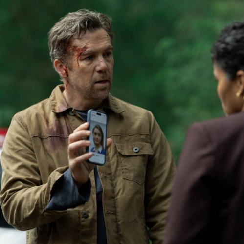 Fotografía de escena en plano medio de Patrick Brammall personificando al detective Ian Ridley. El actor aparece con una herida con sangre en el lado derecho de la frente, vistiendo una chaqueta de pana café sobre una playera oscura. Sostiene frente a él un teléfono celular mostrando la fotografía de una joven en pantalla hacia otra persona de espaldas que viste un saco color guinda.