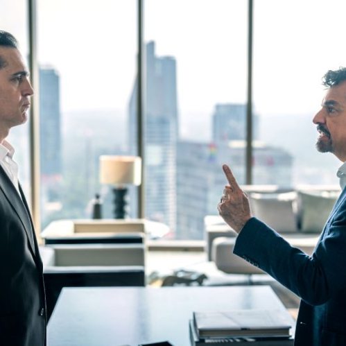Fotografía de escena en plano medio que muestra un enfrentamiento entre Pedro Alonso y Eugenio Derbez en una oficina corporativa. A la izquierda, Pedro Alonso viste un traje negro formal sin corbata y mantiene una postura rígida, mientras que a la derecha Eugenio Derbez, luciendo barba, saco azul y camisa blanca, apunta directamente con el índice hacia el rostro de su interlocutor en señal de reclamo.