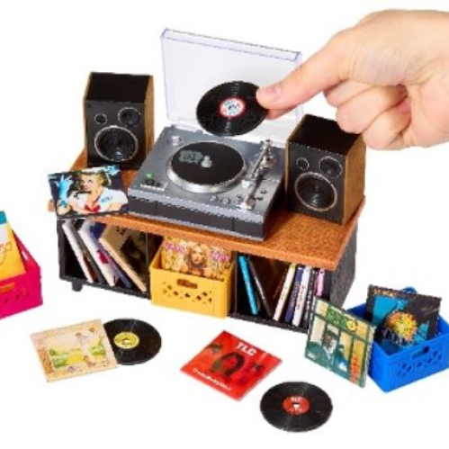 Fotografía en plano cenital sobre fondo blanco de una mano sosteniendo un mini disco de vinilo sobre un pequeño tocadiscos gris funcional con cubierta transparente. A su alrededor se aprecian mini bocinas, un mueble de madera en miniatura, huacales de colores con portadas de álbumes y reproductores a escala de diversos artistas.