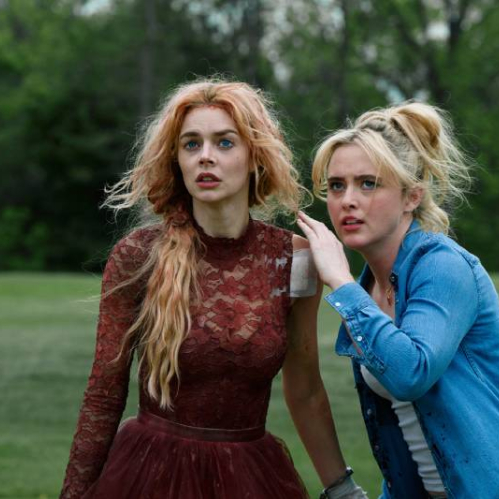 Dos mujeres observan con atención algo fuera de cuadro en un entorno natural, una de ellas con vestido rojo y la otra con camisa azul, ambas mostrando preocupación en su expresión.