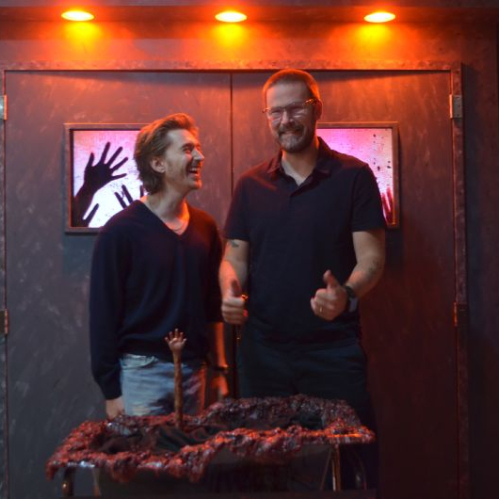 El actor Austin Abrams y el director Zach Cregger posan sonrientes y de pie en un set temático decorado con iluminación roja y elementos de utilería de terror durante la presentación de la película en Ciudad de México.