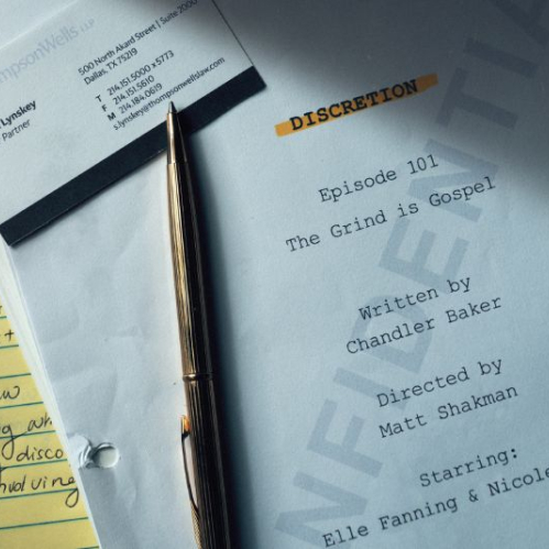 Plano detalle en ángulo picado sobre un escritorio de oficina. Sobre un bloc de notas amarillo se observa el guion del episodio 101 titulado "The Grind is Gospel" de la serie "Discretion", escrito por Chandler Baker y dirigido por Matt Shakman, donde se leen los nombres de Elle Fanning y Nicole Kidman. A la izquierda descansa una tarjeta de presentación del bufete Thompson Wells LLP a nombre de Sharon Lynskey, Senior Partner, junto a un bolígrafo dorado.