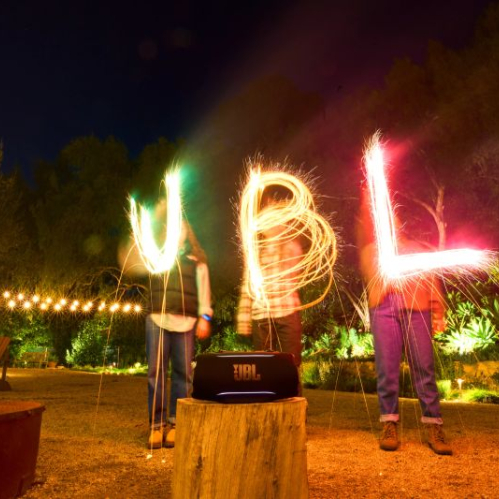 Tres personas sostienen luces de bengala que forman letras luminosas mientras una bocina JBL se muestra en primer plano sobre un tronco.