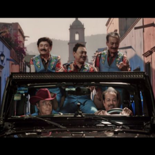Los integrantes de Los Tigres del Norte viajan sonrientes a bordo de un vehículo todoterreno negro a través de una calle empedrada flanqueada por fachadas de colores y una torre de iglesia al fondo.