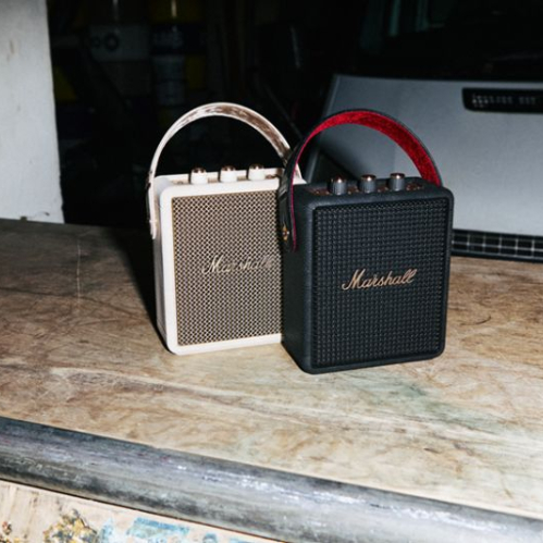 Dos altavoces portátiles Marshall, uno en color crema y otro en negro, colocados sobre una superficie de madera en un entorno industrial.