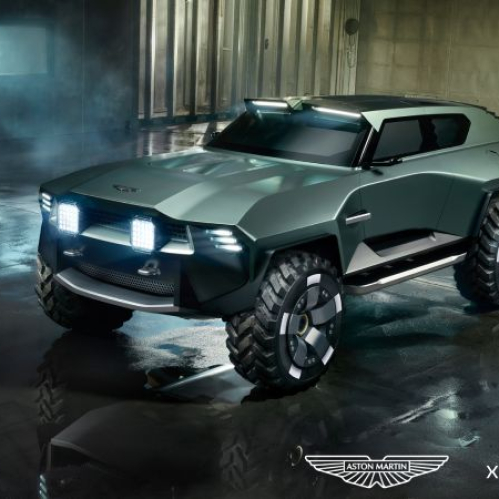Fotografía de estudio que presenta el modelo físico a escala real del SUV táctico de lujo Dreadnought, diseñado por Aston Martin para el videojuego "Call of Duty: Modern Warfare 4". El vehículo, de color verde militar metalizado, se encuentra estacionado sobre un suelo de concreto mojado dentro de un hangar industrial oscuro con tuberías y luces amarillas de fondo. Destaca su imponente frente con faros LED de rejilla brillante, neumáticos todoterreno de gran tamaño con rines de cinco radios y una silueta aerodinámica blindada. En la esquina inferior derecha se aprecian los logotipos oficiales de la colaboración: el emblema alado de Aston Martin al lado del logotipo blanco de "Call of Duty".
