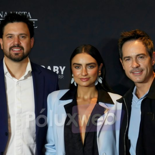 Fotografía de tres personas posando sonrientes de medio cuerpo frente a un fondo negro con letras blancas parciales. De izquierda a derecha aparece un hombre con barba vistiendo traje azul y camisa blanca desabotonada, una mujer al centro con blazer claro y aretes grandes, y un hombre a la derecha con chaqueta oscura y playera polo azul claro.