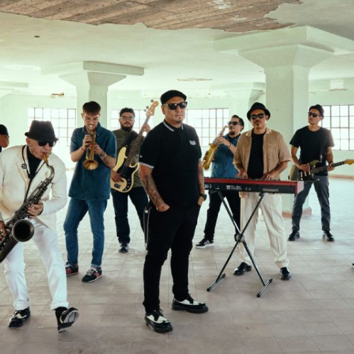 Fotografía a gran escala de la banda de ska mexicano Inspector. En un espacio interior amplio con columnas de concreto y ventanas industriales, los músicos posan portando trajes oscuros, camisas blancas, sombreros estilo pork pie y lentes oscuros. En el centro, el vocalista destaca con un polo oscuro, mientras otros integrantes tocan saxofón, trompeta, teclado y guitarra. La iluminación es natural y el estilo es sobrio y elegante.