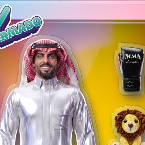 Gráfico promocional de Masad Al-Tamimi representado como un muñeco de acción dentro de una caja de juguete rosa y azul. Viste un thawb tradicional blanco y un keffiyeh rojo y blanco en la cabeza, sonriendo a la cámara. Al lado derecho de la caja se observan accesorios de juguete como un guante de boxeo negro con la inscripción MMA, un pequeño león de peluche tejido y una bufanda doblada.