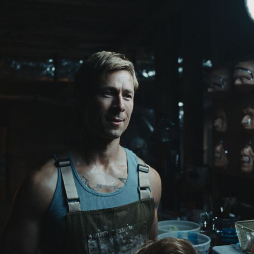 Fotografía en penumbra que muestra al actor Glen Powell caracterizado en un taller o camerino frente a un soporte con prótesis faciales y máscaras.