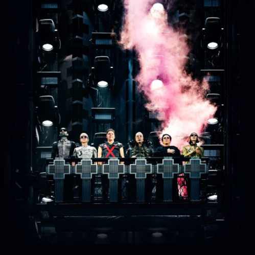 Los seis integrantes de la banda Rammstein se encuentran alineados sobre una plataforma metálica elevada en medio de una escenografía industrial iluminada, mientras una densa nube de humo rosa asciende detrás de ellos.