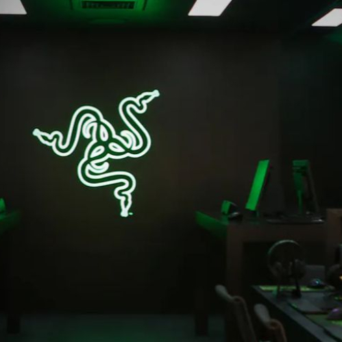 Sala de gaming con estaciones de cómputo y un logotipo verde iluminado de Razer en la pared.
