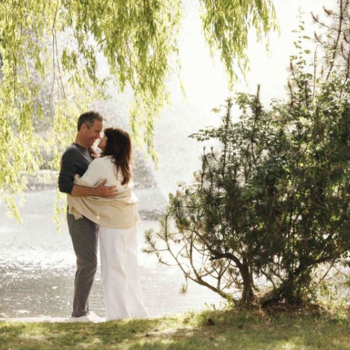 Una pareja de adultos abraza sonriente junto a un cuerpo de agua rodeado de árboles y vegetación bajo la luz del sol.