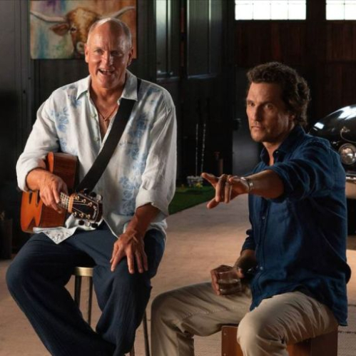 Dos personas conversan en un espacio tipo estudio o garaje, con una guitarra, un automóvil clásico y elementos decorativos alrededor.