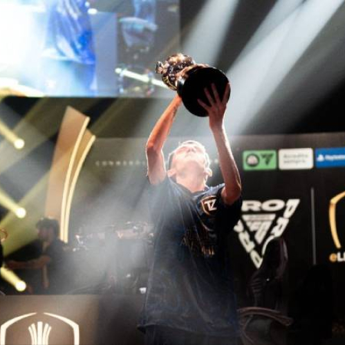 Fotografía de un jugador levantando un trofeo en un escenario iluminado, rodeado de pantallas y elementos visuales propios de un torneo profesional de esports.