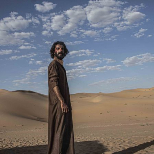 Un hombre con barba y túnica café observa de perfil a la distancia parado sobre la arena en medio del desierto bajo un cielo despejado con nubes dispersas.