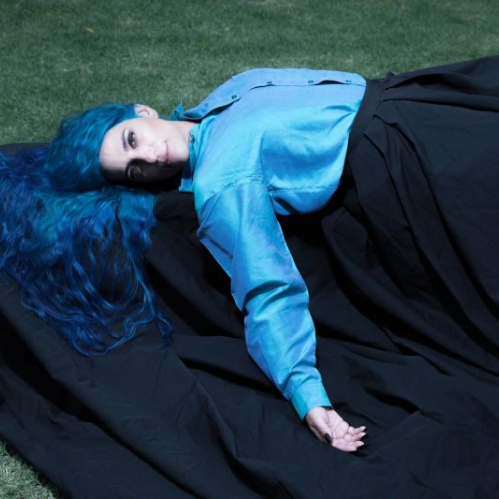 Fotografía de la cantautora Sofía Rei recostada en el césped vistiendo una camisa azul turquesa y falda negra, luciendo su característico cabello largo de color azul.